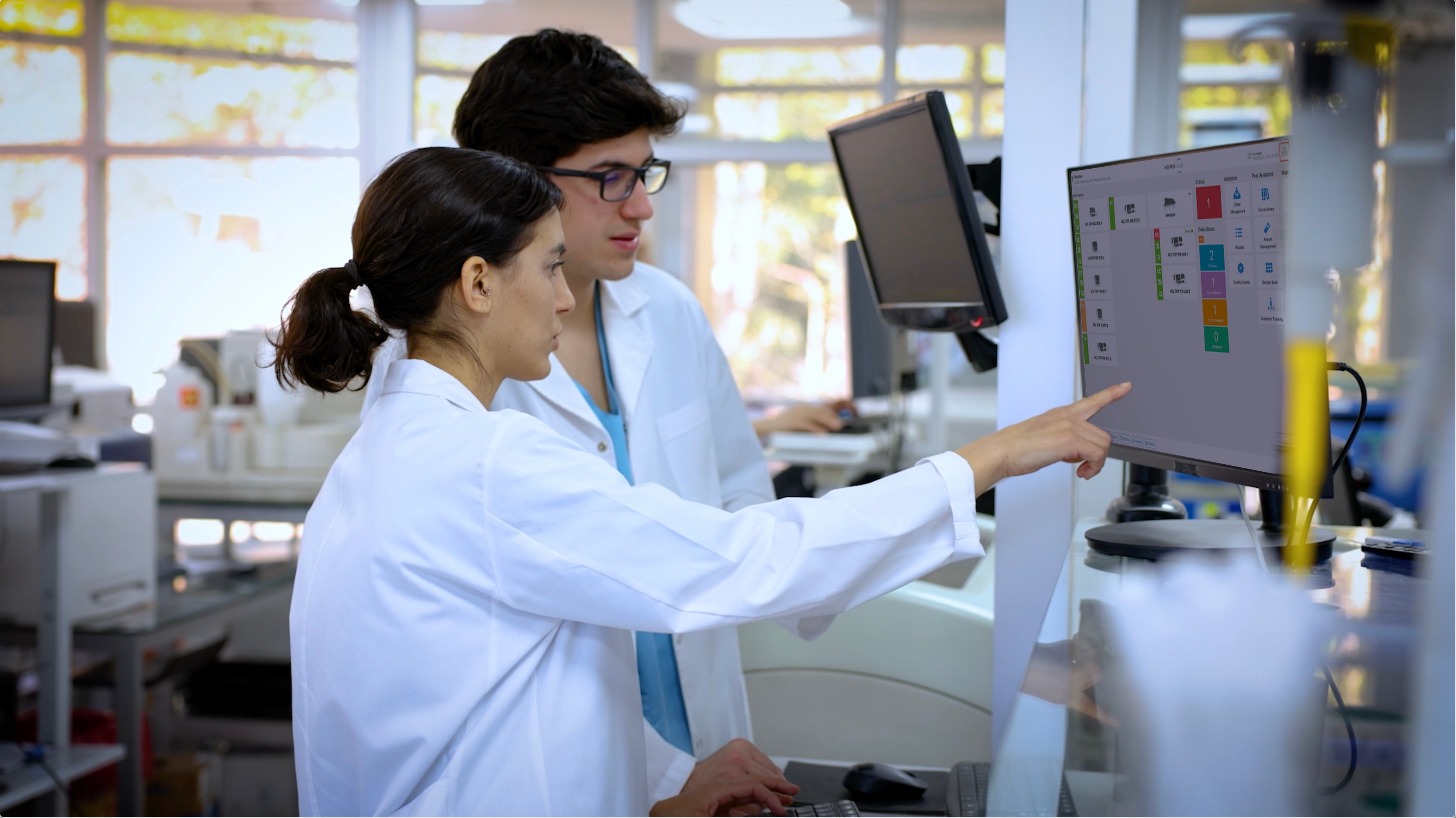 Photo of a man and woman in a laboratory setting both wearing white lab coats. They are looking at a monitor that shows the HemoHub interface and the woman is pointing to something on the monitor.