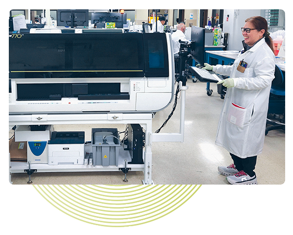 Women standing in a medical laboratory wearing a long, white lab coat and smiling. She is working on the keybard that is attached to the ACL TOP 700 analyzer. Light green circles are placed artistically in the background.
