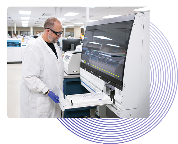 Man standing in a medical laboratory wearing a long, white lab coat and safety goggles. He is loading cuvettes into  HemoCell automation cell. Light blue circles are placed artistically in the background.