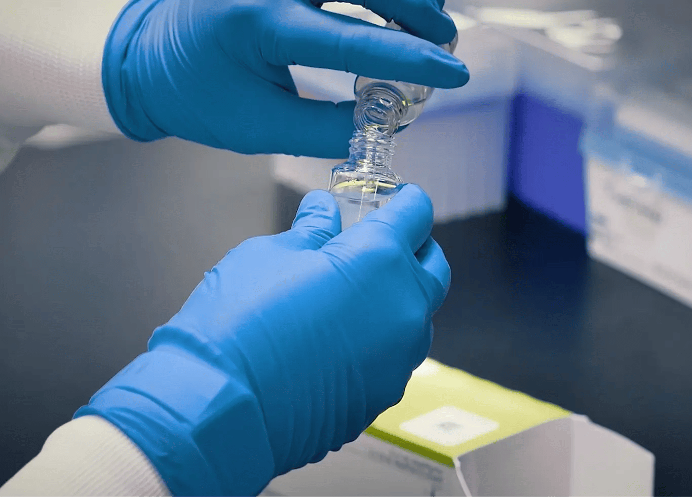 Photo of a person's hands wearing blue laboratory gloves and pouring a liquid reagent into another bottle. The branded green top of the HemosIL Reagent box can be seen in the background.